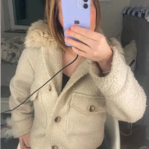 Chelsea teddyjacka - Supermysig beige teddyjacka med fluffig fuskpäls-krage och stora fickor framtill. Jackan är croppad och har knappar hela vägen. Perfekt för dig som vill ha en trendig och varm look under höst och vinter.