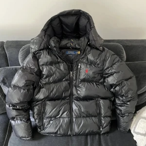 Pufferjacka från Polo Ralph Lauren - Säljer en fet pufferjacka från Polo Ralph Lauren med ikonisk röd logga på bröstet. Jackan har hög krage, avtagbar huva och flera dragkedjefickor. Finns i både svart och mörkblå, perfekt för kalla dagar. Materialet är glansigt och känns riktigt lyxigt.