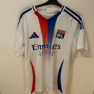 Lyon hemma tröja 24/25 cherki - Säljer en officiell Olympique Lyonnais matchtröja från Adidas med Cherki 18 på ryggen. Tröjan är vit med röda och blå detaljer, klubbmärke på bröstet och klassiska Adidas-ränder på axlarna. Slim fit-modell i lätt och ventilerande material.