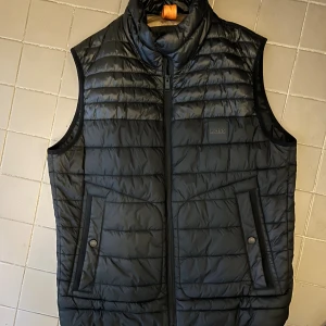 Svart väst från Hugo BOSS - Snygg svart väst från BOSS med quiltad design och dragkedja framtill. Västen har två fickor med knappstängning och en hög krage. Insidan är fodrad i beige och orange detaljer. Perfekt för lager-på-lager-stil under kyliga dagar. Ny pris runt 2000, knappt använd.