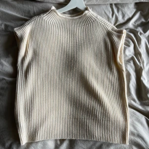 Beige ribbad stickad väst - Säljer en beige stickad väst med ribbad struktur och hög hals. Västen är ärmlös och har en avslappnad passform, perfekt att slänga över en skjorta eller t-shirt för en chill look.