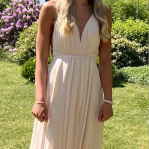 Ljusbeige långklänning med slits - Superfin ljusbeige långklänning med djup v-ringning, öppen rygg och tunna axelband som korsas bak. Klänningen har en hög slits framtill och faller snyggt i ett lätt, flowy material. Perfekt balklänning. Banden kan nytas olika hårt så storleken regleras (xs-m). Finns två små hål i undertyget men syns inte vid användning då de hänger tyger över, (går o sy ihop hålen eller klippa av tyget där de är)