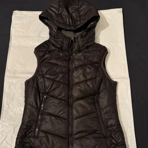 Svart dunjacka - Svart dunväst från H&M Sport med quiltad design och avtagbar huva. Västen har dragkedja framtill och två fickor med dragkedja. Perfekt för lager-på-lager och funkar till både chill och aktivitet.