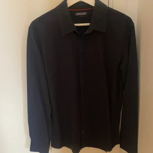 Mörkblå skjorta från Tommy Hilfiger - Stilren svart skjorta från Tommy Hilfiger med klassisk krage och diskret logga på bröstet. Skjortan har långa ärmar och knäppning framtill. Perfekt för dig som gillar enkel och snygg design med en touch av premiumkänsla.