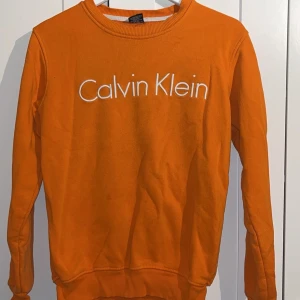 Orange sweatshirt från Calvin Klein - Snygg orange sweatshirt från Calvin Klein med vit broderad logga framtill. Tröjan har rund halsringning, långa ärmar och ribbade muddar vid ärmslut och nederkant. Perfekt för dig som vill sticka ut med färg och stil.