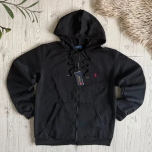 Svart hoodie från Polo Ralph Lauren - Svart hoodie från Polo Ralph Lauren med dragkedja framtill och klassisk huva med snörning. Tröjan har en liten röd broderad logga på bröstet och två fickor framtill. Tillverkad i mjuk bomull och har långa ärmar för en chill look.
