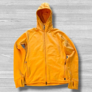 Gul fleecejacka med huva från Houdini - Snygg gul fleecejacka från Houdini i storlek XL. Jackan har huva, två sidofickor med dragkedja och tumhål vid ärmslut. Materialet är mjuk polyesterblandning som håller dig varm och bekväm. Perfekt för chill dagar eller när du är ute och rör på dig.