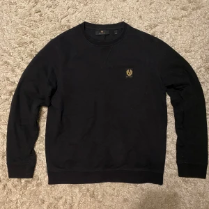Svart sweatshirt från Belstaff - Svart sweatshirt från Belstaff med rund hals och lång ärm. Tröjan har en snygg patch med Belstaff-loggan på bröstet. Tillverkad i mjuk bomull med lite elastan i muddar för extra komfort. Perfekt för chill dagar och enkel att matcha med jeans.