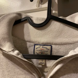 Beige half zip tröja från Next Supply - Säljer en beige tröja från Next Supply med half zip och hög krage. Tröjan är i bomullsmix och har en avslappnad passform med ribbade muddar vid ärmslut och nederkant. Perfekt för lager-på-lager under kyliga dagar.