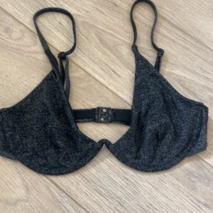 Svart glittrig bikiniöverdel - Snygg svart bikiniöverdel med glittrigt tyg och bygel för extra stöd. Justerbara axelband och klassisk knäppning bak. Perfekt för dig som vill sticka ut på stranden eller vid poolen.