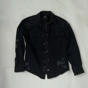 Svart overshirt från Zara med slitningar - Svart overshirt från Zara med coola slitningar och rå kant nedtill. Skjortan har två bröstfickor med knappar, klassisk krage och tryckknappar framtill. Materialet är kraftig bomull som ger en schysst streetkänsla. Perfekt för dig som gillar en edgy stil.