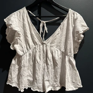 Vit blus med volangärm från Lager157 - Supersöt vit blus från Lager157 med korta volangärmar och v-ringning. Blusen har knyt i ryggen och är tillverkad i ett lätt, krinklat bomullstyg som ger en chill och luftig känsla. Perfekt för varma dagar och enkel att styla med jeans eller kjol.