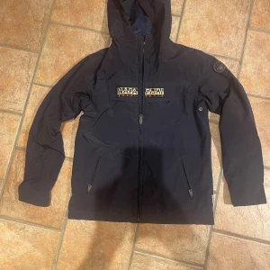 vindjacka Napapijri - Snygg mörkblå vindjacka från Napapijri med huva och dragkedja framtill. Jackan har meshfoder och stort Napapijri-märke på bröstet. Nypris 1800