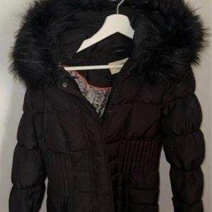 Svart pufferjacka från Roco Baroco - Svart pufferjacka från Roco Baroco med fluffig fuskpäls på huvan och snyggt paisleymönstrat innerfoder. Jackan har dragkedja framtill och markerad midja för en schysst siluett. Perfekt för kalla dagar när du vill vara både varm och stilren.