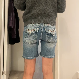 Ljusblå jeansshorts med broderade fickor - Säljer ett par ljusblå jeansshorts från Miss Me med snygga slitningar och broderade bakfickor med knappar. Klassisk femficksmodell, lågmidjade och i stretchig bomull. Perfekta för sommaren och ger en cool, avslappnad vibe.