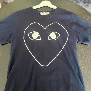 Svart t-shirt Comme des Garçons Play - Svart t-shirt från Comme des Garçons Play med ikoniskt stort hjärta med ögon tryckt i vitt på bröstet. Klassisk passform, rund hals och korta ärmar. Tillverkad i mjuk bomull som känns skön mot huden. Perfekt statement-plagg för dig som gillar streetstyle.