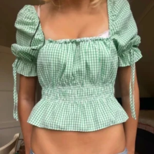 Grön rutig croppad blus med puffärm - Superfin grön och vit rutig blus från H&M med croppad passform och puffiga korta ärmar. Blusen har smock i midjan, volangkant nedtill och snörning vid ärmslut. Perfekt för dig som gillar romantisk och trendig stil.