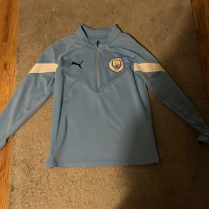 Manchester City blå träningshoodie Puma - Snygg ljusblå träningshoodie från Puma med Manchester City-logga på bröstet och svart Puma-märke. Tröjan har lång ärm, half zip och vita detaljer på ärmarna. Tillverkad i DryCell-polyester som andas och håller dig torr under fotbollsträningen.