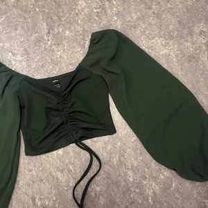 Mörkgrön croppad topp från SHEIN - Säljer en mörkgrön croppad topp från SHEIN med långa puffärmar och snygg snörning framtill. Toppen har v-ringad hals och är tillverkad i mjukt syntetmaterial. Perfekt för dig som gillar trendiga detaljer och vill sticka ut lite extra.
