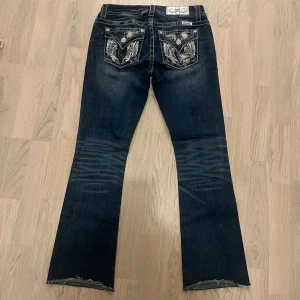 Miss Me jeans med paljettdetaljer - Mörkblå jeans från Miss Me med bootcut-modell och snygga slitningar. Bakfickorna har broderier och paljetter i silver, samt dekorativa knappar. Jeansen har kontrastsömmar och råa benslut för en edgy look.