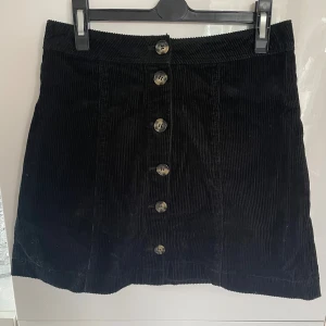 Svart manchesterkjol med knappar - Snygg svart kortkjol i manchester med raka linjer och knappar hela vägen framtill. Kjolen har en klassisk, enkel design och passar perfekt till en cool vardagslook. Tjockt, strukturerat tyg som ger en trendig känsla.