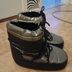 Silvriga moon boots med snörning - Moon boots i metallic silver och svart med tjock, vadderad ovandel och svarta snören runtom. Snyggt futuristiska och perfekta för kalla dagar. Platt sula och rund tå, insida i mjukt material för extra komfort.