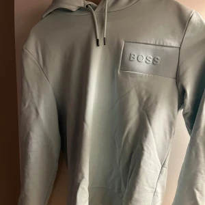 Ljusblå hoodie från Hugo Boss - Snygg ljusblå hoodie från Hugo Boss med stilren design och diskret BOSS-logga på bröstet. Tröjan har huva med snörning. Perfekt för en chill och trendig look. Stl M/S, har två små hål