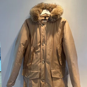 Beige Woolrich Arctic Parka med päls - Säljer en beige Woolrich Arctic Parka med fluffig päls på huvan. Jackan har långa ärmar, stora fickor med knappar och klassisk parkamodell. Fylld med dun för extra värme och tillverkad i slitstark polyester. Perfekt för kalla dagar.