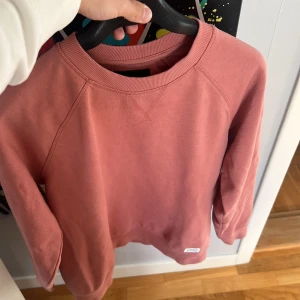 Rosa sweatshirt i bomull - Säljer en enkel och snygg rosa sweatshirt med rund hals och ribbade muddar. Tröjan har långärmade raglanärmar och en liten patch vid nederkanten. Perfekt för chill dagar och passar till jeans eller mjukisbyxor.