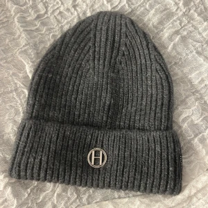 Grå ribbad mössa från Hollies - Snygg grå mössa från Hollies med ribbad stickning och uppvikt kant. Metallmärke med H-logga framtill och diskret svart etikett på sidan. 