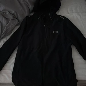 Svart vindjacka från Under Armour - Svart vindjacka från Under Armour med huva och reflekterande detaljer. Jackan har dragkedja framtill och logga på bröstet. Perfekt för löpning eller utomhusaktiviteter. Tillverkad i lätt polyester för maximal rörelsefrihet.