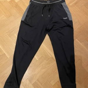 Svarta mjukisbyxor från BOSS XL - Svarta mjukisbyxor från BOSS med gråa detaljer vid fickorna, logga på vänster sida och mudd vid benet. Byxorna har resår och snörning i midjan för skön passform. Perfekta för chill eller träning, tillverkade i mjukt material. Använd fåtal gånger och i bra skick.