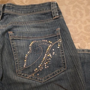 Blå jeans med strass från Lindex, stl 36 - Snygga blå jeans från Lindex Jeans med strassdetaljer i silver och guld på bakfickan, men några har ramlat. Jeansen är lite utsvängd men men som raka ben. Jeansen är i stretchig denim som sitter skönt och har en stilren look. Om man tycker om lite oversized och loose fit passar den bra för curvy S annars ger mer tajt fit för curvy M. Kolla på sista bild för veta om skick!                                     Skriv gärna om du vill fler bilder! Står inte för frakt och tar ingen retur 🩷