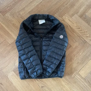 Moncler jacka  - Svart dunjacka från Moncler med quiltad design och klassisk logga på ärmen. Jackan har dragkedja framtill . Insidan har en cool serietidningsetikett. Perfekt för dig som vill ha en snygg och lätt jacka med bra kvalitet. Pris kan diskuteras 