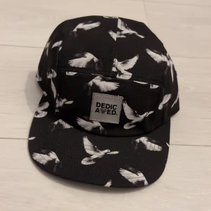 Svart keps med fågelmönster från Dedicated - Svart keps från Dedicated med vitt fågelmönster över hela kepsen. Klassisk fempanelsdesign med justerbar rem bak och tydlig logotyp framtill. Tillverkad i bomull och designad i Sverige. Perfekt för dig som vill sticka ut med en unik accessoar.