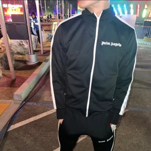 Palm Angels svart hoodie med vita ränder - Svart hoodie från Palm Angels med vita ränder längs ärmarna och vit dragkedja framtill. Tröjan har hög krage och logotypen broderad på bröstet. Snygg streetwear-look, perfekt för dig som gillar trendiga märken och sportig stil. Den är copy 