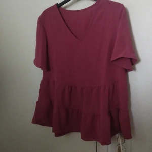 Rosa/lila volangblus från SHEIN - Säljer en rosa/Lila  blus från SHEIN i storlek M. Toppen har v-ringning, korta ärmar och flera volanglager nedtill som ger en flowy och chill vibe. Perfekt för dig som gillar en lite lösare passform och vill ha något bekvämt men ändå snyggt.