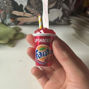 Fanta Strawberry Lip Smacker läppbalsam - Supergulligt läppbalsam från Lip Smacker i samarbete med Fanta Strawberry. Kommer i en röd plastförpackning som ser ut som en Fanta-bägare med sugrör. Själva läppbalsamet är rosa och har en rundad, äggformad design som är enkel att applicera.