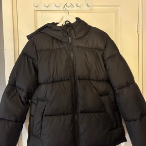 Svart pufferjacka med huva - Säljer en svart pufferjacka med huva och snörning. Jackan har en rejäl dragkedja framtill och två sidofickor. Perfekt för kalla dagar och har en snygg, oversized look som är riktigt trendig just nu.