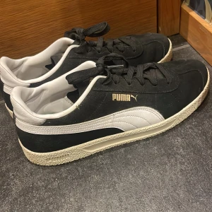 Svarta och vita Puma sneakers strl 41 - Klassiska Puma sneakers i svart mocka med vita detaljer och logga i guld på sidan. Skorna har vit sula med struktur, rund tå och snörning. Snygg kontrast mellan svart och vitt, samt ikonisk Puma-formstripe i läder på sidan. Perfekta för en streetwear-look.