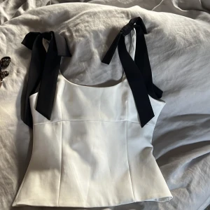 Vit linnetopp med svarta rosetter Zara - Supersnygg vit linnetopp från Zara med breda svarta knytband på axlarna som ger en trendig look. Toppen har en figurnära passform. Perfekt för dig som vill sticka ut med stilrena detaljer.