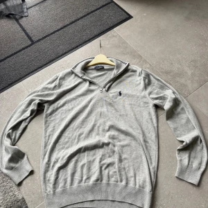 Grå half zip tröja från Polo Ralph Lauren - Snygg grå långärmad tröja från Polo Ralph Lauren med half zip och klassisk broderad logga på bröstet. Tröjan är stickad i mjuk bomull och har ribbade muddar vid ärmslut och nederkant. Perfekt för dig som gillar stilren och enkel design.