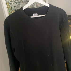Svart sweatshirt från CP Company - Svart långärmad sweatshirt från CP Company med klassisk rund hals och ikonisk linsdetalj på ärmen. Tröjan har en clean look och är tillverkad i mjuk bomull som är skön att bära. Perfekt för dig som gillar streetwear och stilrena plagg.