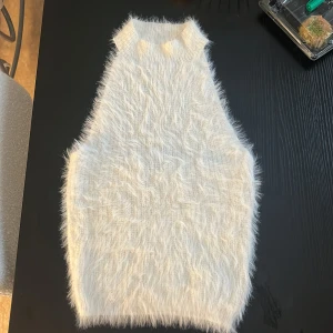 Vit fluffig ärmlös stickad topp - Supermjuk och fluffig vit stickad topp med hög hals och ärmlös design. Toppen har en cool, lite croppad passform och är perfekt för att sticka ut med sin unika textur. Passar snyggt till både jeans och kjol för en trendig look.