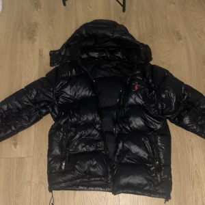 Svart pufferjacka från Polo Ralph Lauren - Säljer en svart pufferjacka från Polo Ralph Lauren med huva och två stora fickor med dragkedja. Jackan har en röd Polo-logga på bröstet och är riktigt fluffig och varm. Perfekt för kalla dagar och har en snygg, glansig finish.                         Finns 2 defekter att ena dragkedjan är lite sönder