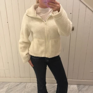 Vit teddyjacka från 157 XS - Mysig vit teddyjacka från 157 i storlek XS. Jackan har fluffig yta, dragkedja framtill och två fickor. Perfekt för dig som gillar en chill och varm stil. Passformen är normal och jackan har långa ärmar. Snygg att matcha med jeans eller leggings.