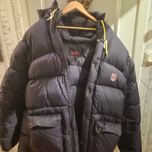 Svart dunjacka från Fjällräven storlek XL - Säljer en svart, riktigt fet dunjacka från Fjällräven med huva och gula snören. Jackan har stora fickor med knappar, dragkedja och klassisk Fjällräven-logga på bröstet. Perfekt för kalla dagar och har en schysst oversized vibe. XL Finns i Hässelby 