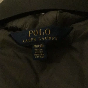 Svart lång dunjacka från Ralph Lauren - Lång svart dunjacka från Polo Ralph Lauren med huva och quiltad design. Jackan har en klassisk pufferstil och är perfekt för kalla dagar. Broderad logga på insidan och dragkedja framtill. Snygg och tidlös modell som håller dig varm hela vintern.