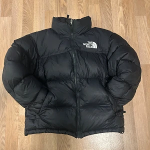 Svart pufferjacka från The North Face - Säljer en svart pufferjacka från The North Face med fet logga på bröstet och ryggen. Jackan har hög krage, dragkedja framtill och är riktigt tjock och fluffig. Perfekt för kalla dagar och har innerficka med dragkedja. Stilsäker och klassisk modell.