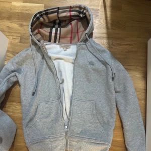 Burberry hoodie/kofta - Grå hoodiejacka från Burberry med klassiskt rutigt foder i beige, svart och rött. Jackan har dragkedja, snörning i huvan och broderad Burberry-logga på bröstet. Tillverkad i mjuk bomull- och polyesterblandning, perfekt för en chill men stilren look. Storlek M, passar även smal med en normal passform.